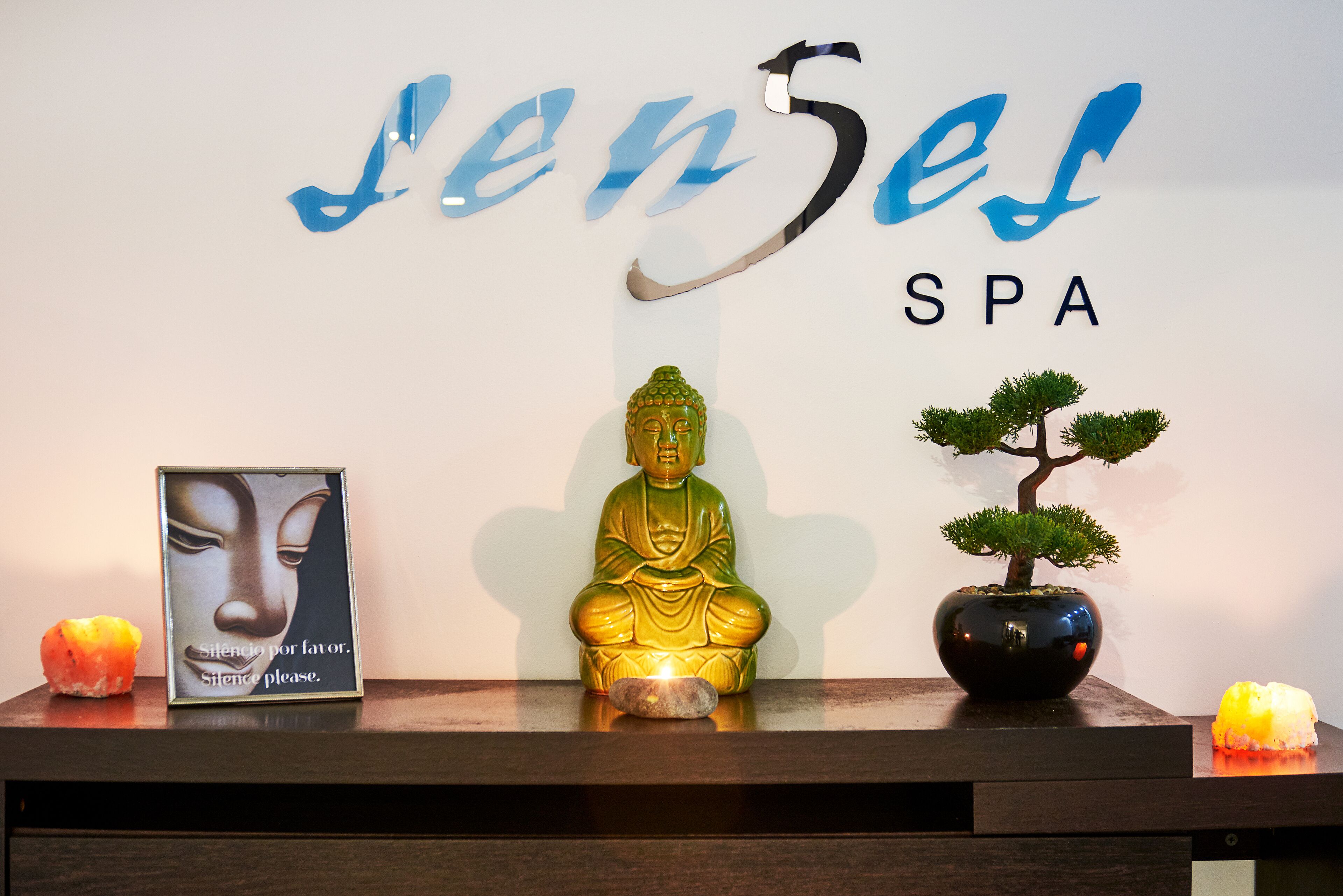 Een receptiebalie met een Boeddhabeeld, een ingelijste foto, een bonsaiboom en een bordje met de tekst 'Senses SPA'.