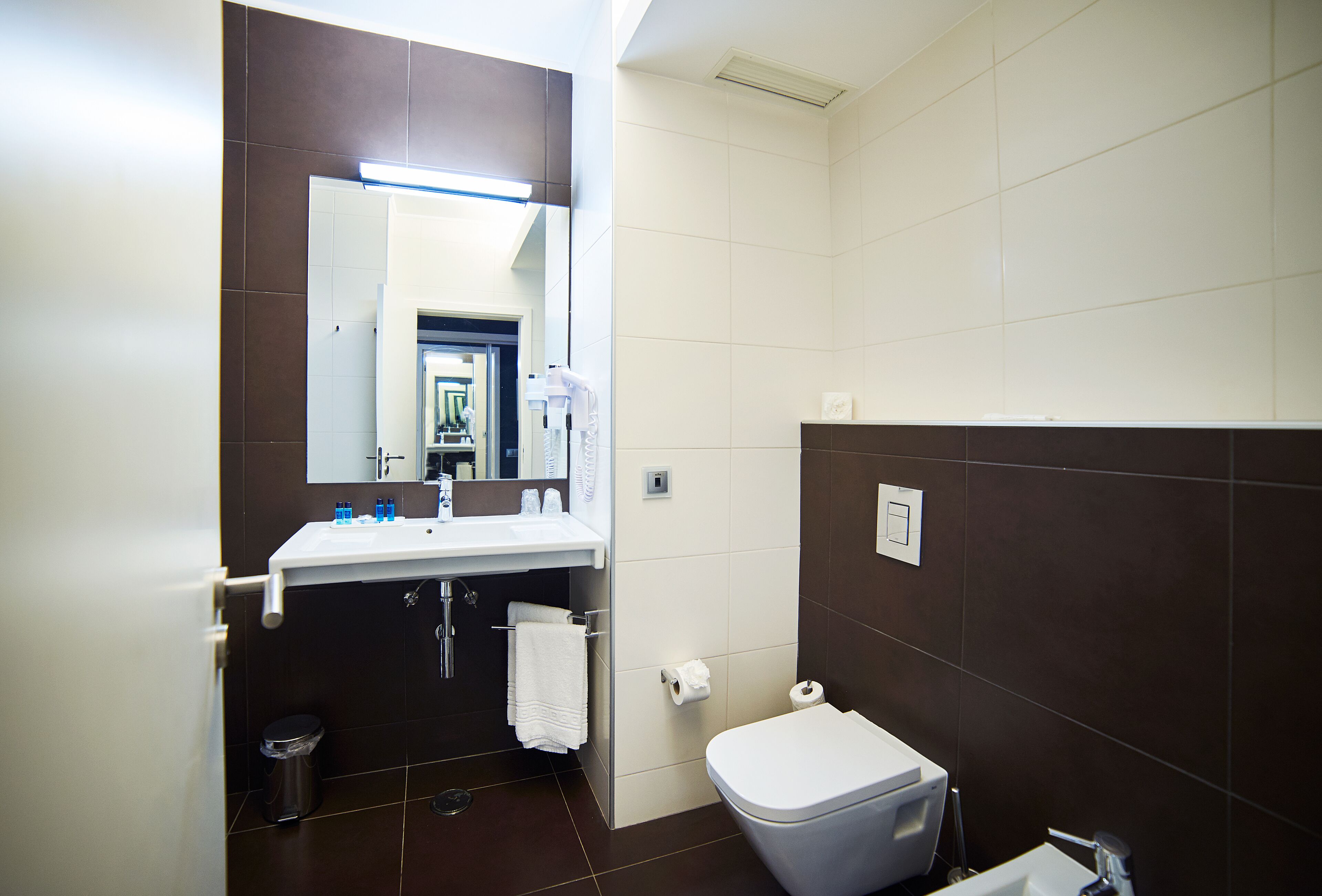 A modern bathroom with a white sink, dark brown tiles, a white toilet, and a mirror above the sink.