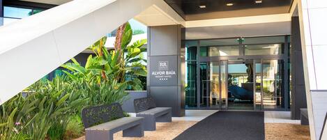 A modern hotel entrance with a glass door, a staircase, and outdoor seating.