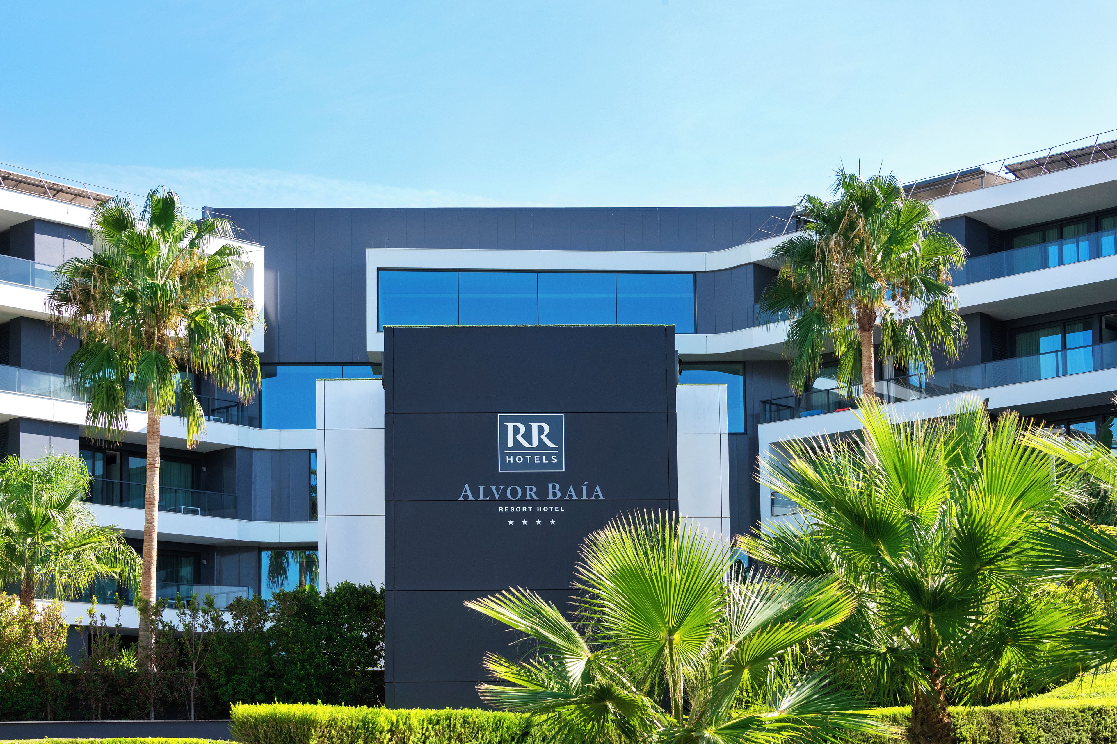 Un moderno edificio de hotel con un letrero llamativo para 'Alvor Baía Resort Hotel' y una vista despejada de la vegetación circundante.