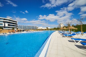 Una gran piscina con agua azul, rodeada de sillas de descanso y sombrillas, con edificios modernos al fondo.