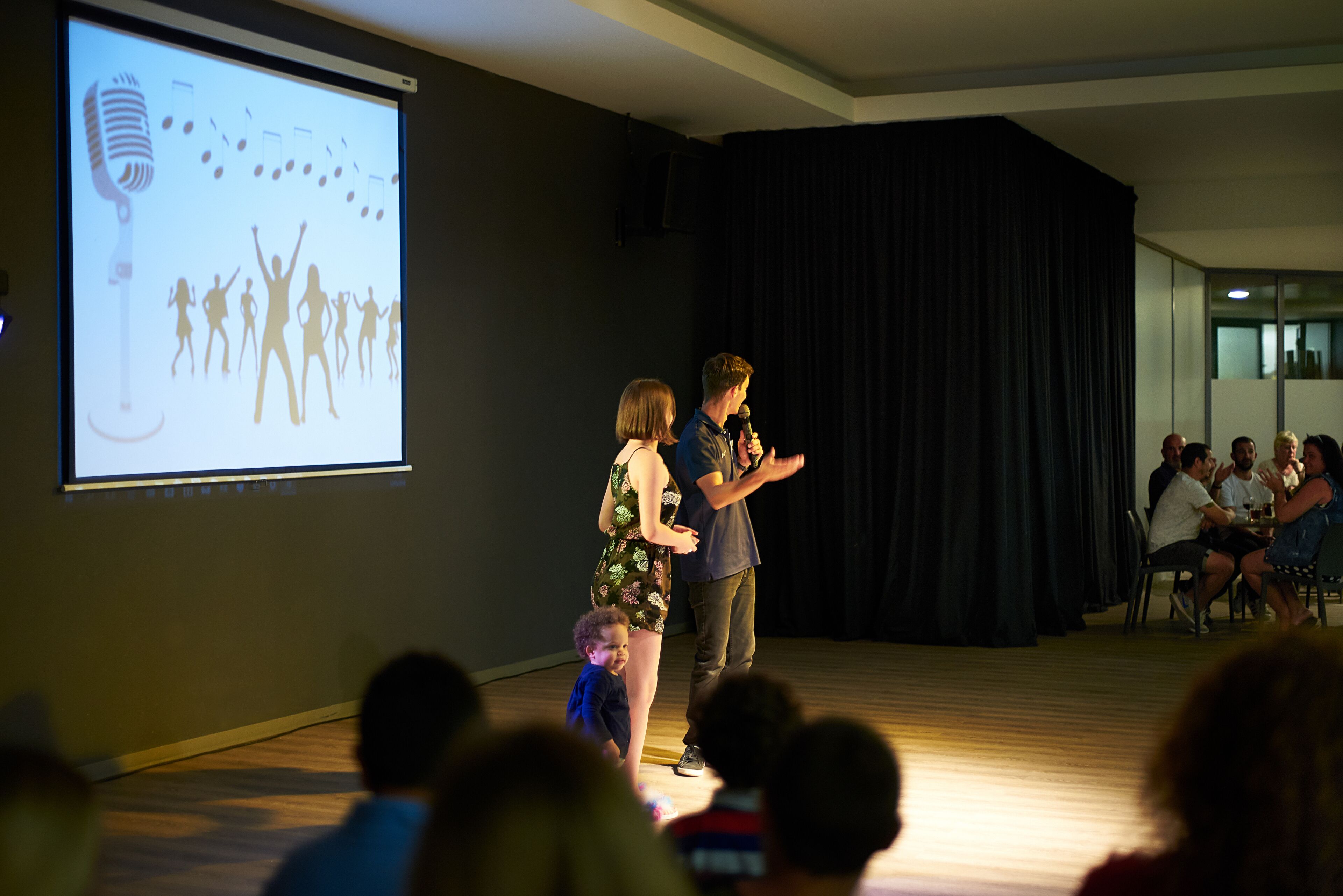 Una presentación en una sala con un escenario, una pantalla que muestra un micrófono y figuras bailando, y una audiencia sentada en mesas.
