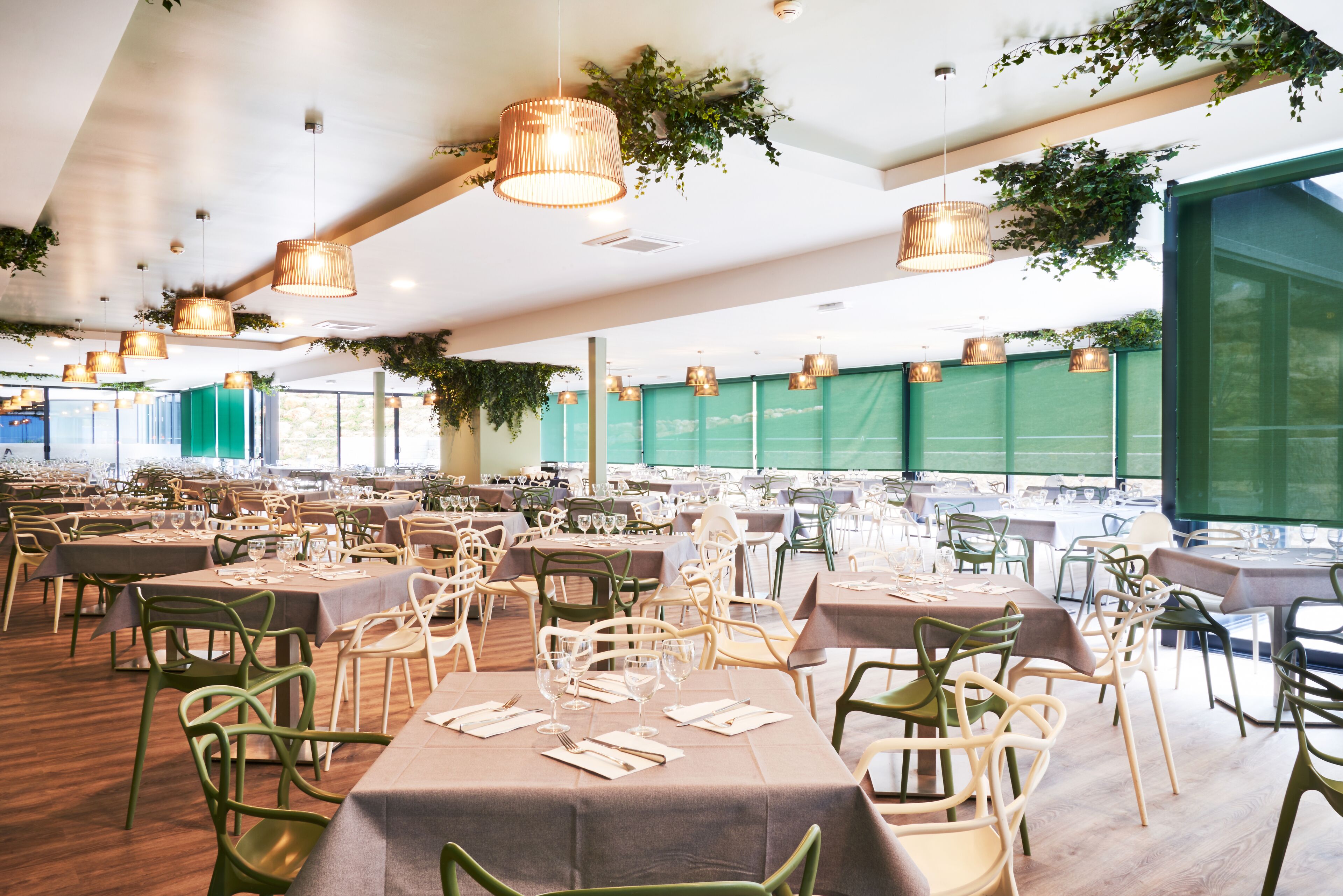 Un vaste espace dédié à la salle à manger, avec des tables dressées pour le repas, entourées de chaises vertes et agrémenté de plantes suspendues.