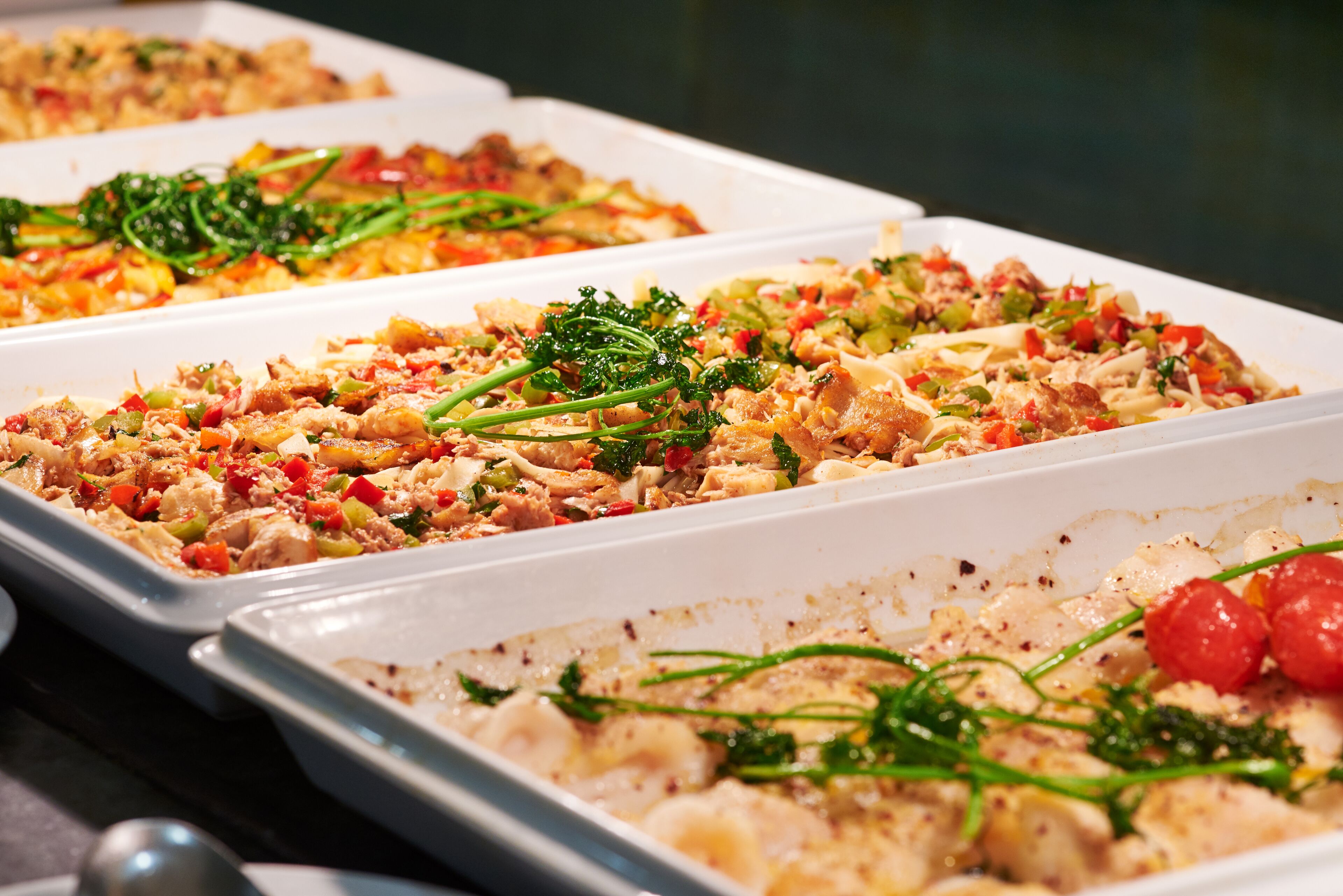Een buffet met diverse gerechten, waaronder pasta met groenten en vlees, gepresenteerd op witte schalen.
