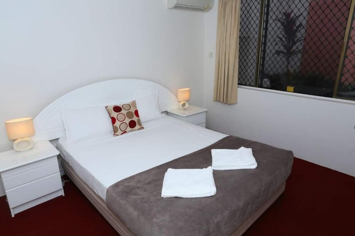 Una cama bien hecha con sábanas blancas, un edredón marrón, dos almohadas blancas y dos toallas dobladas sobre la cama.