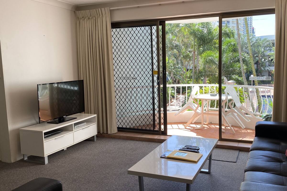 Una sala de estar con televisor, sofá, mesa de centro y puerta corrediza de vidrio que da a un área exterior con piscina y sillas.