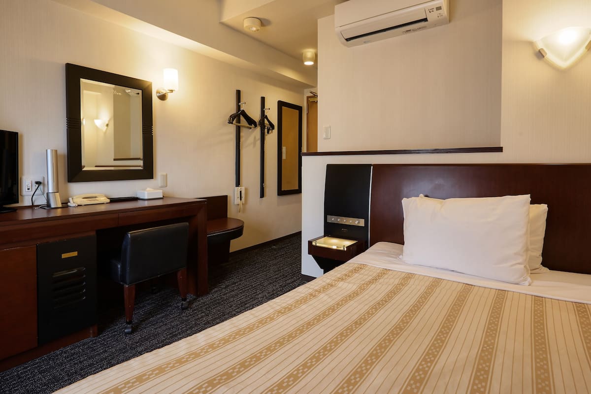 A hotel room with a bed, a desk, a chair, a mirror, a wardrobe, and an air conditioning unit.