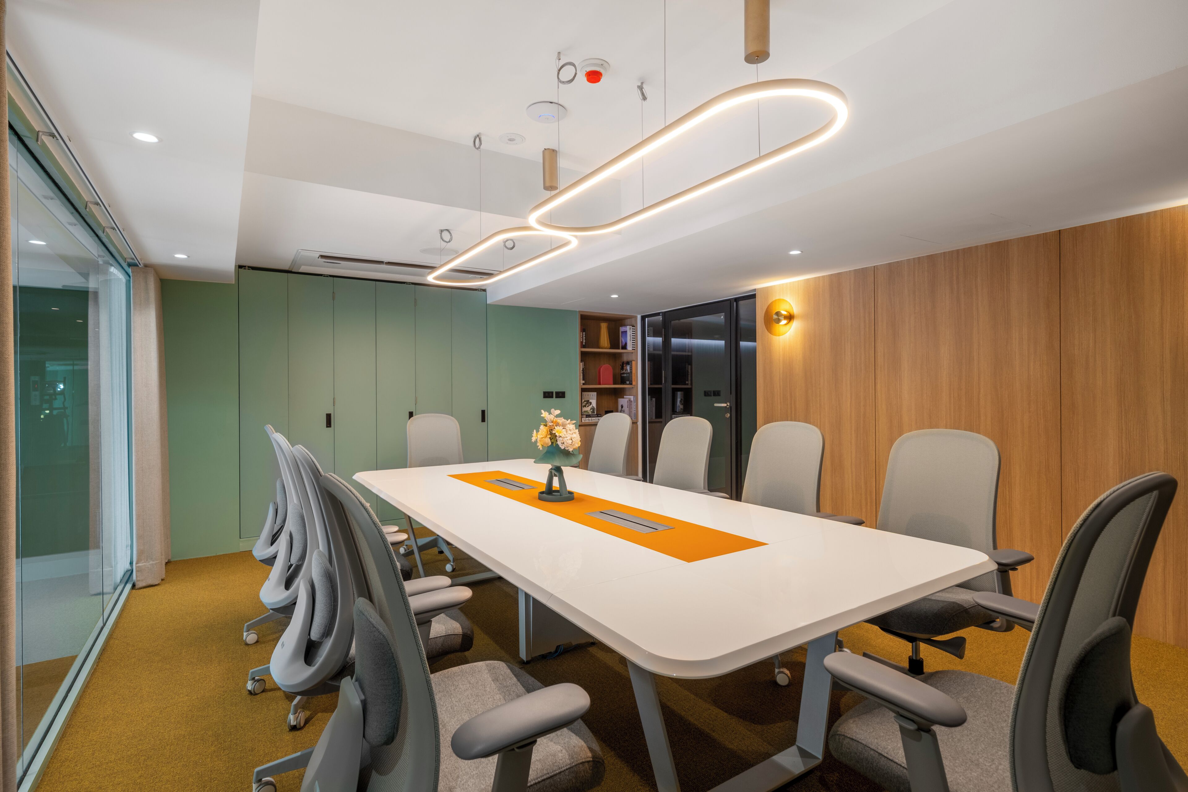 A conference room with a white table, grey chairs, a glass wall, and a wooden panel wall.