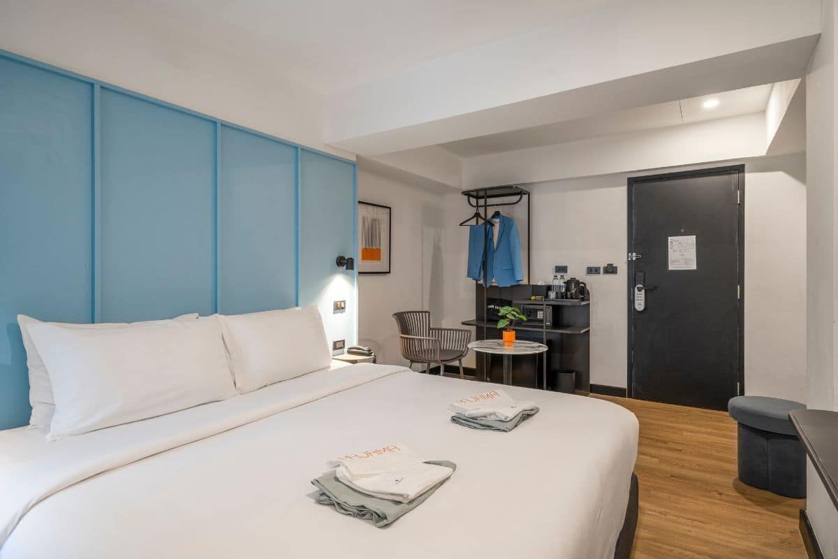 A hotel room with a large bed, a desk with a chair, a small table with a plant, and a wardrobe.