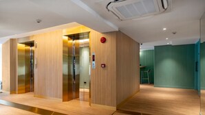 A modern elevator with wooden paneling and a metallic finish, a red emergency button, and a small stool in a hallway with wooden flooring and green walls. - Myhotel CMYK Ratchada (Bangkok)