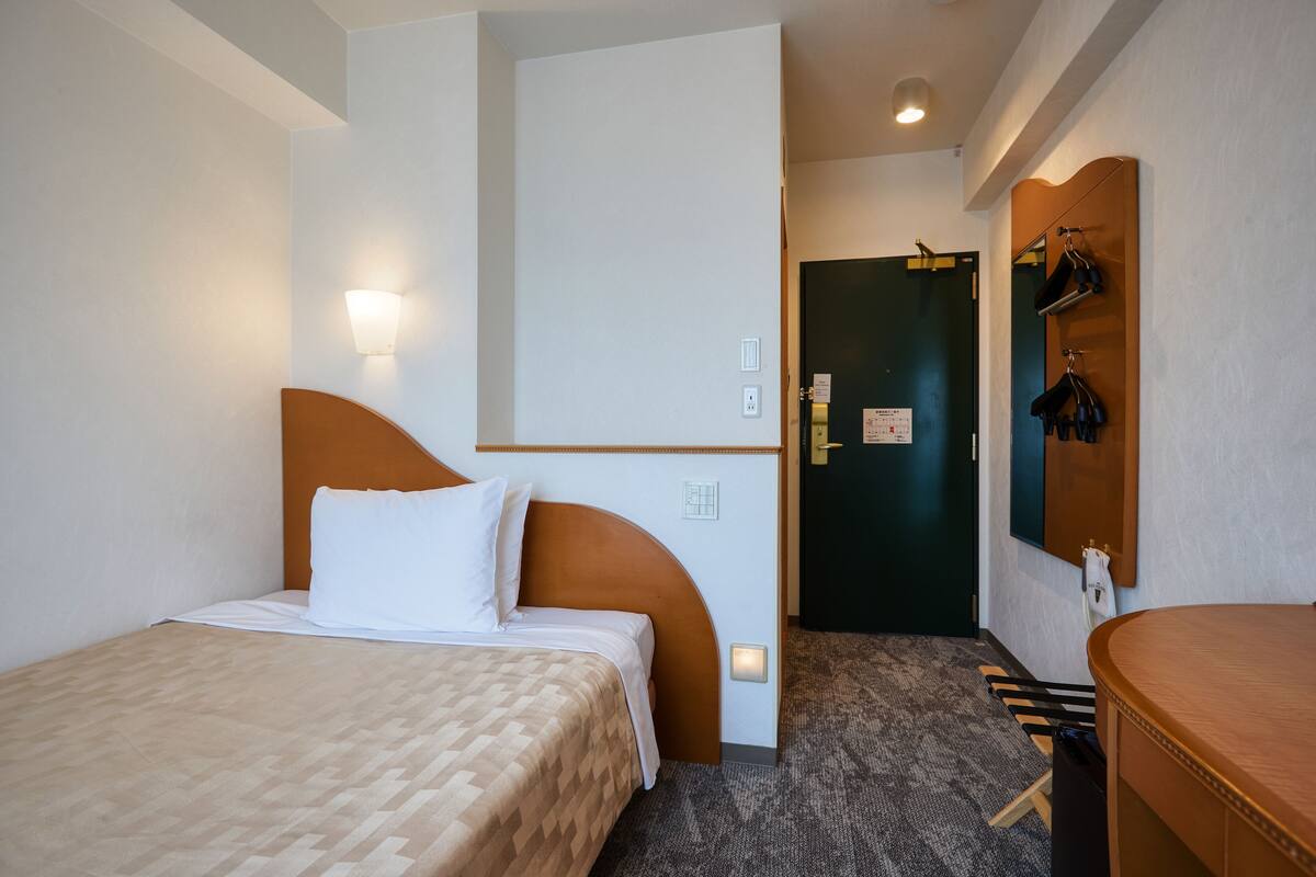 Quarto de hotel com cama, mesa de cabeceira, espelho e porta para outro espaço.