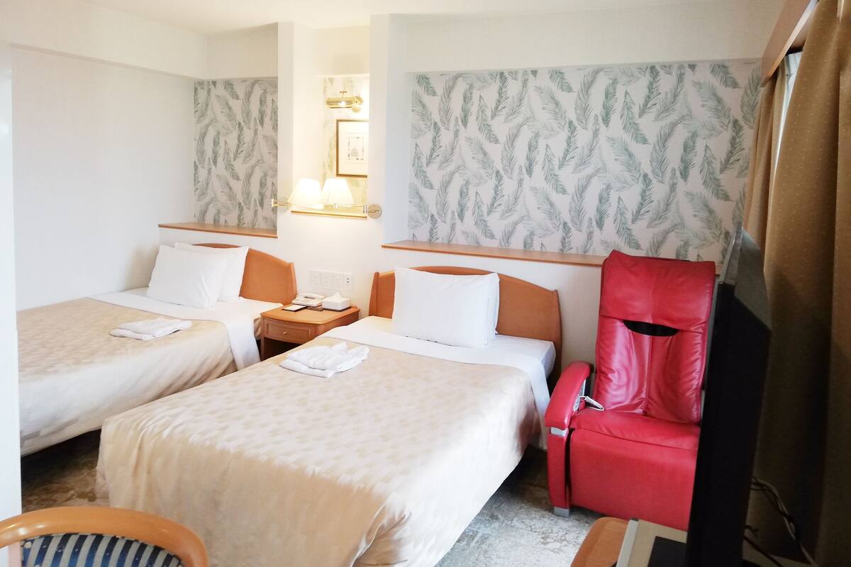 Quarto de hotel com duas camas, uma cadeira vermelha e uma mesa de cabeceira com abajur.