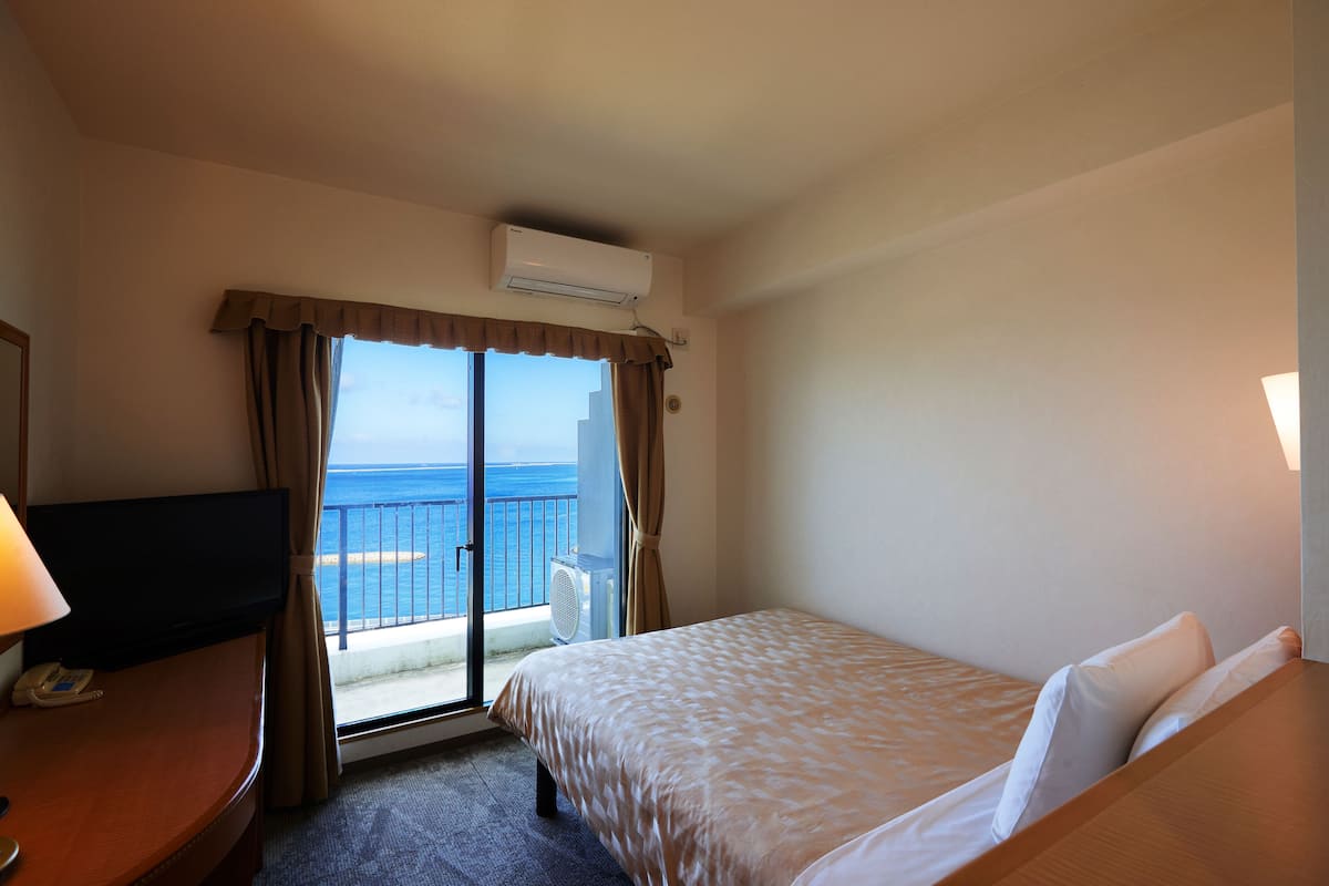 Quarto de hotel com cama, escrivaninha, cadeira e vista para o oceano através de uma porta de varanda.