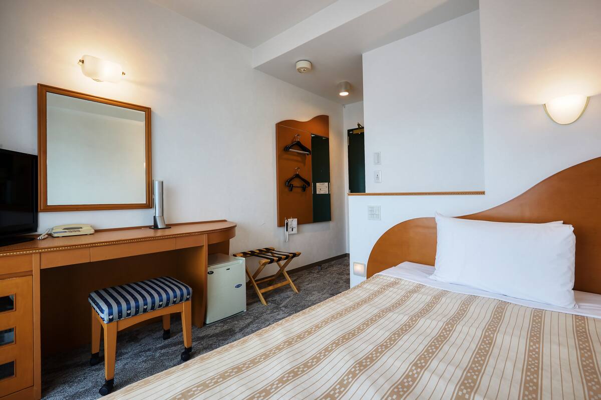 Quarto de hotel com cama, escrivaninha com cadeira, um banquinho pequeno, espelho e guarda-roupa.