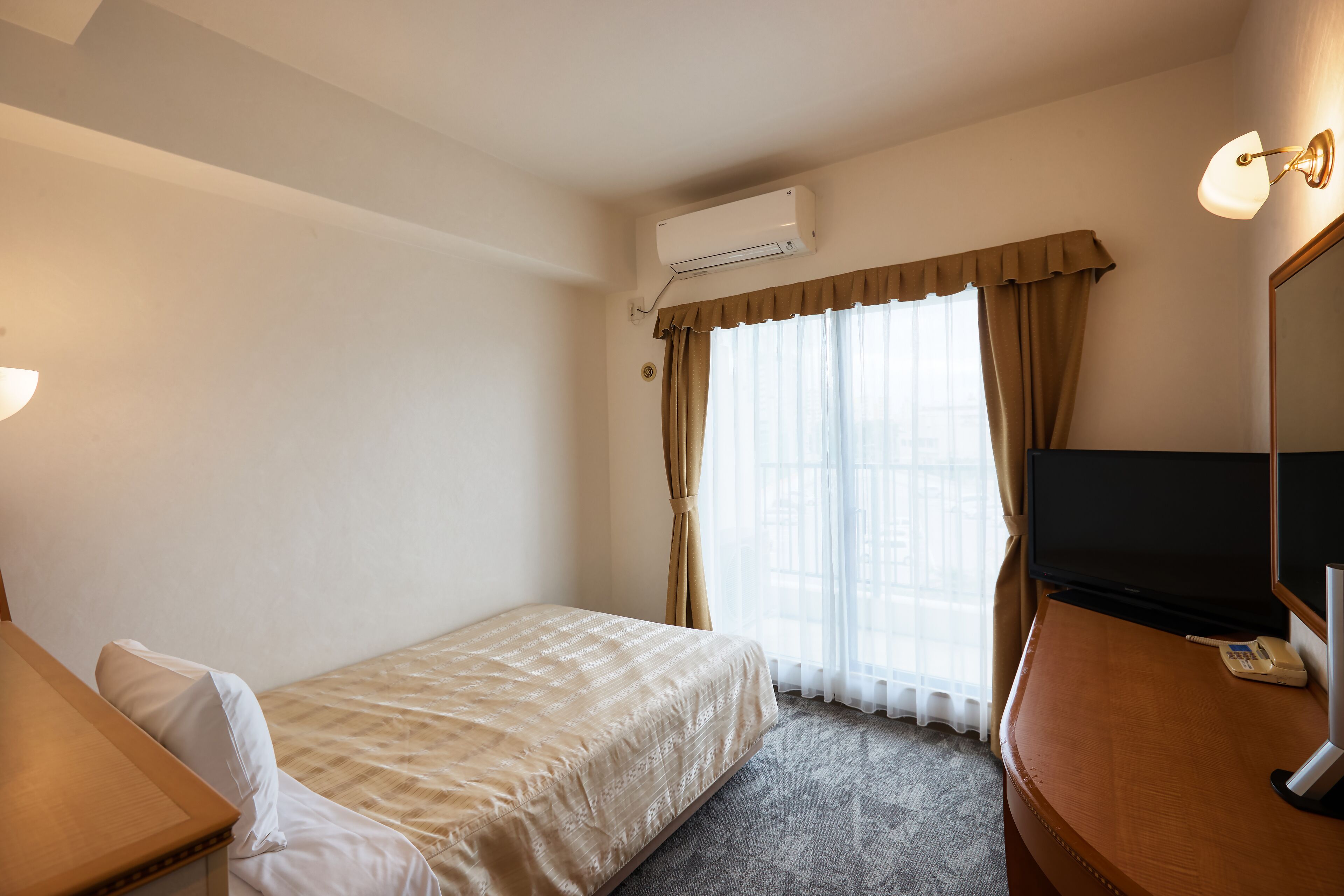 A hotel room with a bed, a desk, a TV, and an air conditioning unit.