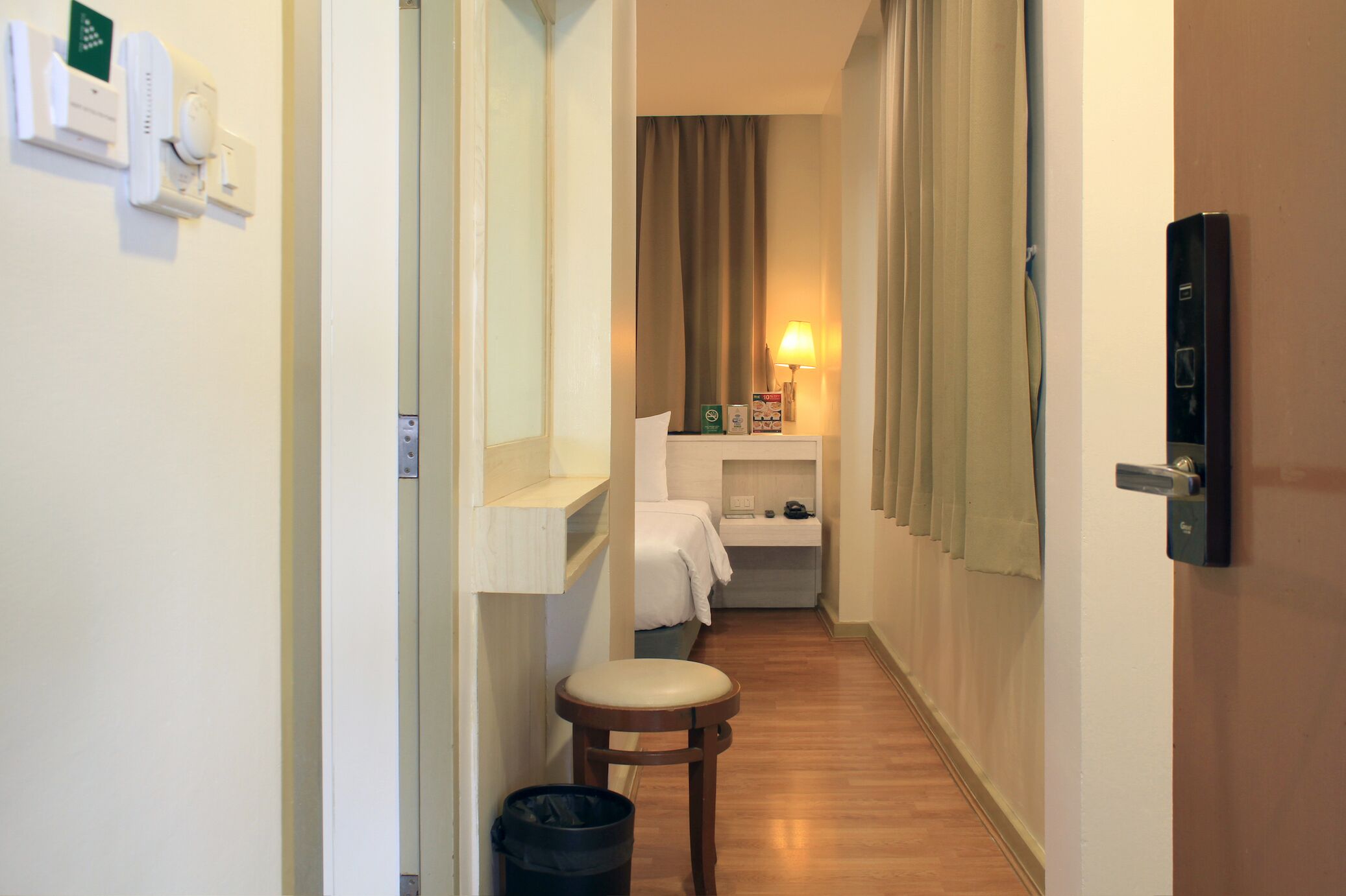 A narrow hotel hallway with a wooden floor, a small stool, a trash bin, a bed with a white cover, a nightstand with a lamp, and a door with a lock.
