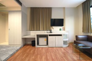 Ein modernes Hotelzimmer mit Holzboden, einem an der Wand befestigten Flachbildfernseher, einem kleinen Kühlschrank, einem Glastisch und einem Ledersofa.