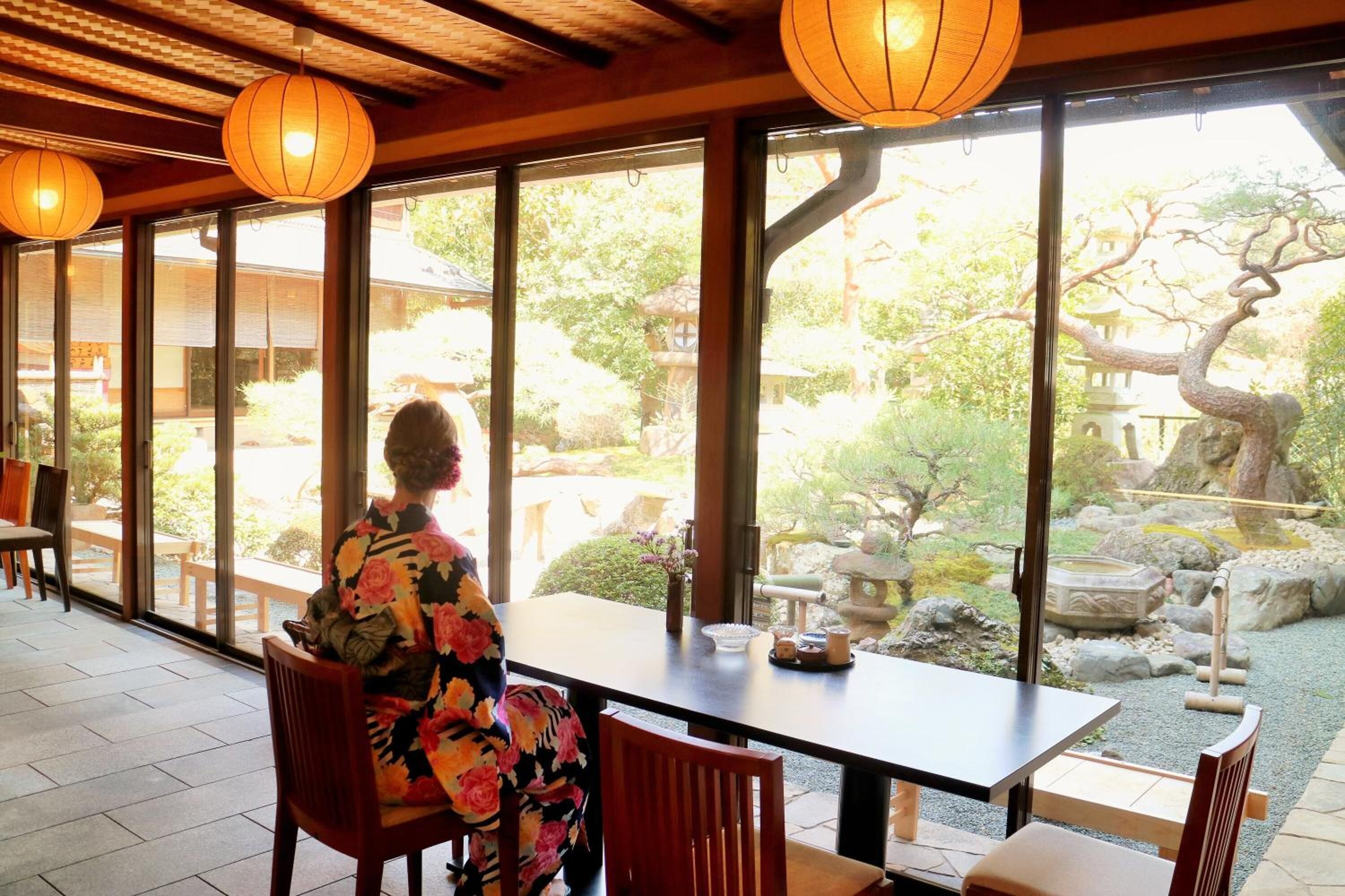 A person seated at a table with a view of a garden through large windows.