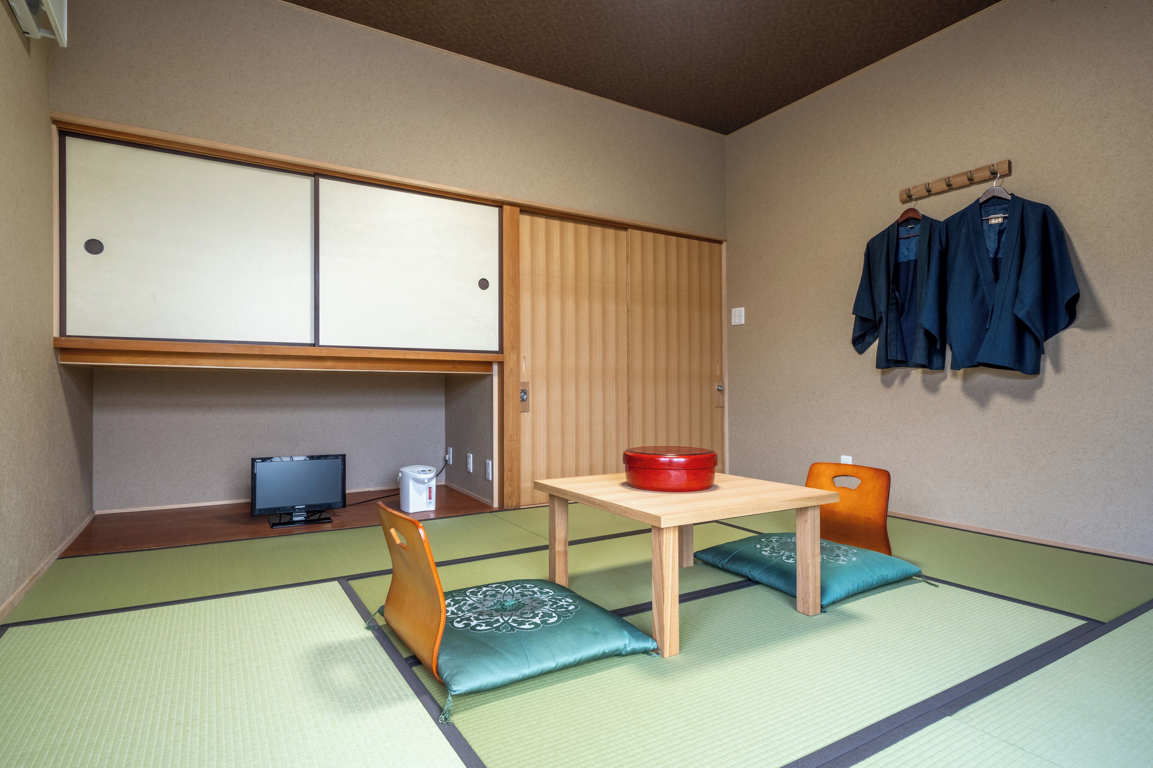Um quarto japonês tradicional com tatames, uma mesa baixa com uma tigela vermelha, uma televisão e um armário de madeira.