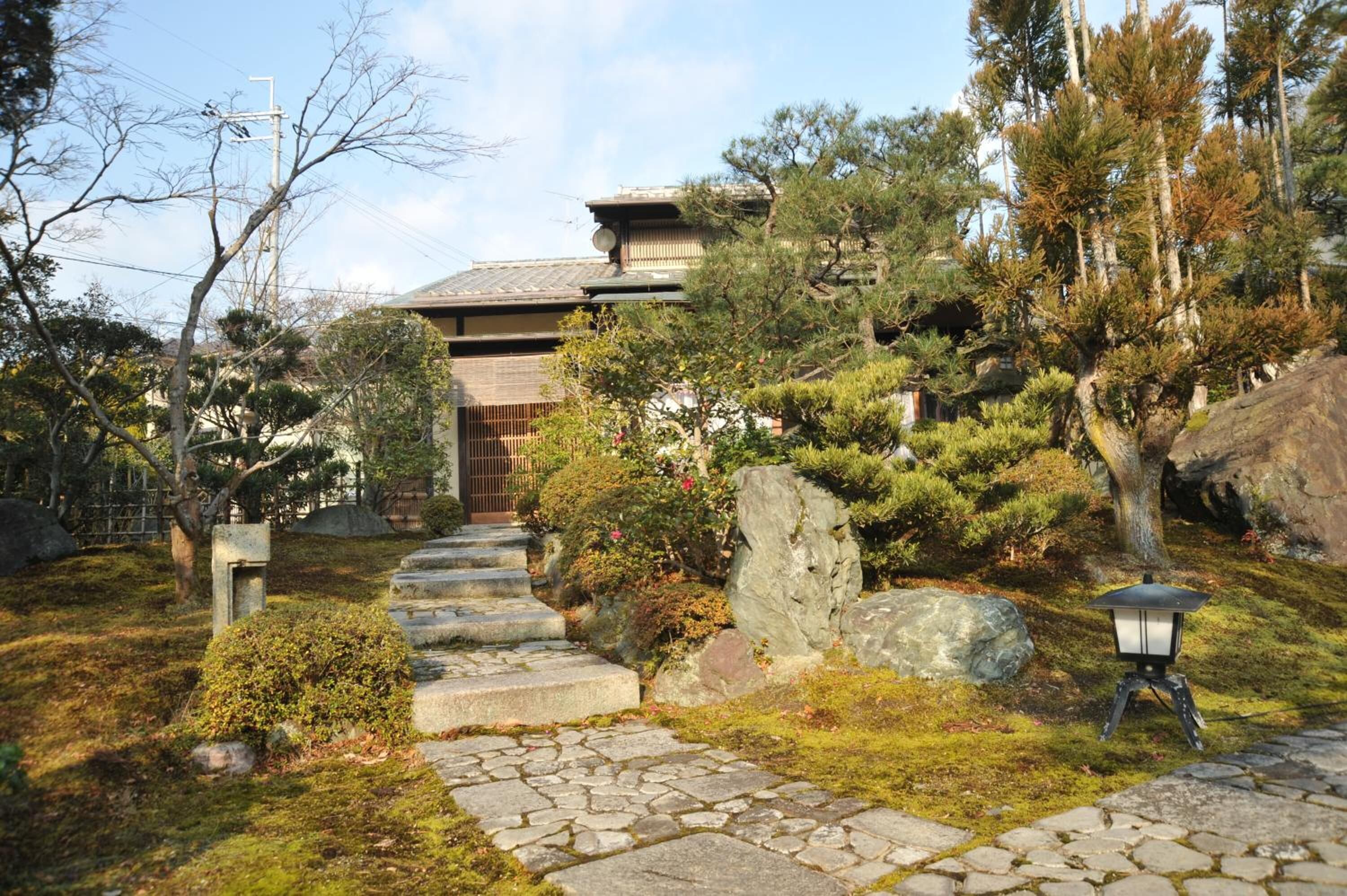A traditional Japanese building with a stone pathway leading to the entrance, surrounded by lush greenery and rocks.