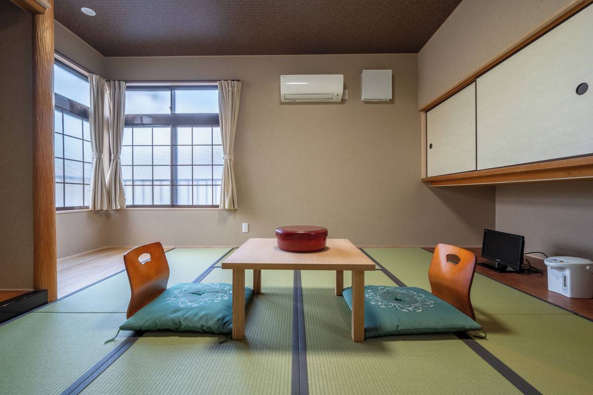 A traditional Japanese room with tatami flooring, a low table with a red bowl, two wooden chairs, a television, and a cabinet.