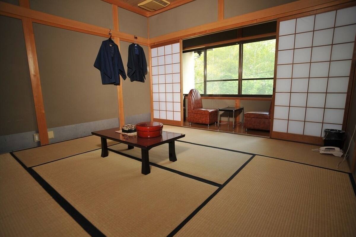 A tatami mat room with a low table, two hanging clothes, and a view of the outdoors through a sliding door.