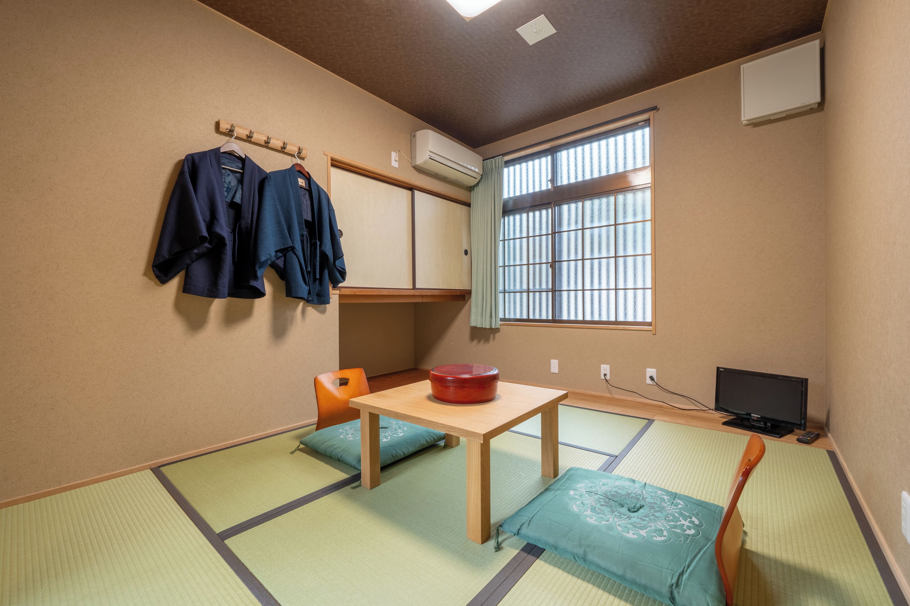 A room with tatami flooring, a wooden table, a red bowl, a television, and a window with blinds.