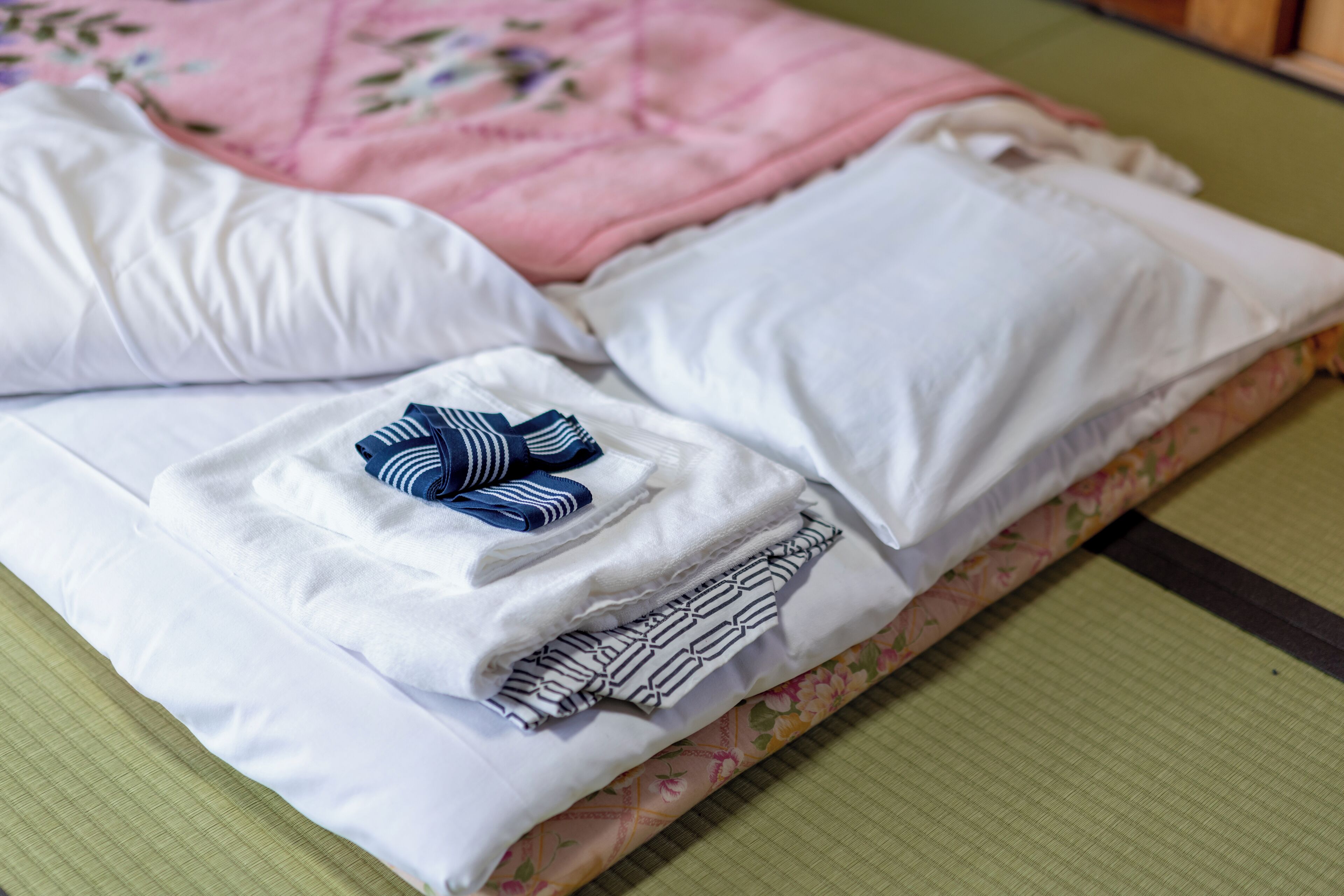 A neatly made bed with white pillows and folded blankets, a floral-patterned bedspread, and a green mat at the foot of the bed.