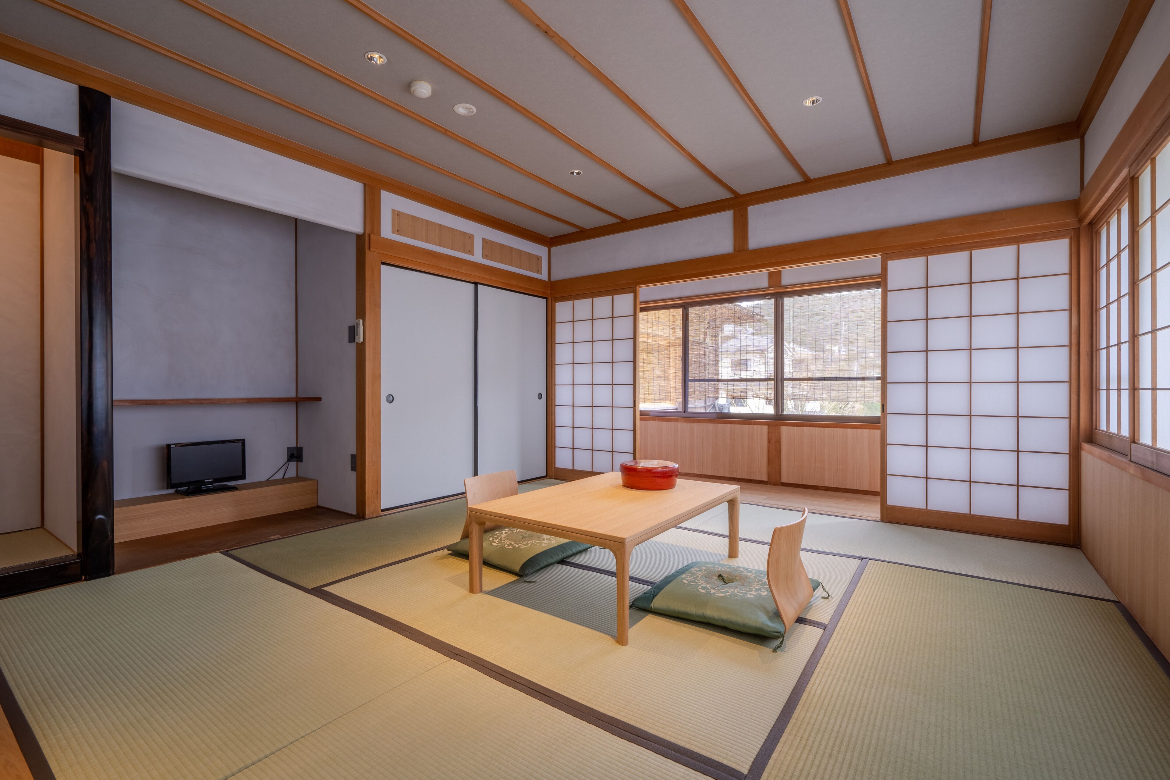 Une pièce de style japonais traditionnel, avec un sol recouvert de tatamis, une table basse avec des coussins, des portes coulissantes et un téléviseur posé sur une étagère.