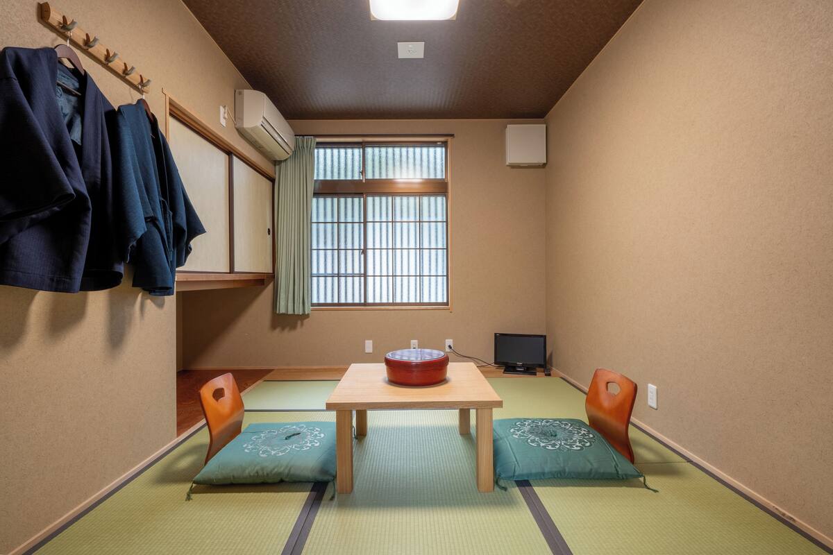 A traditional Japanese room with tatami flooring, a wooden table with a red bowl, two cushions, a television, and a window with vertical blinds.