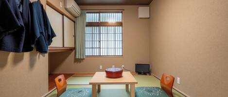 A traditional Japanese room with tatami flooring, a wooden table with a red bowl, two cushions, a television, and a window with vertical blinds.
