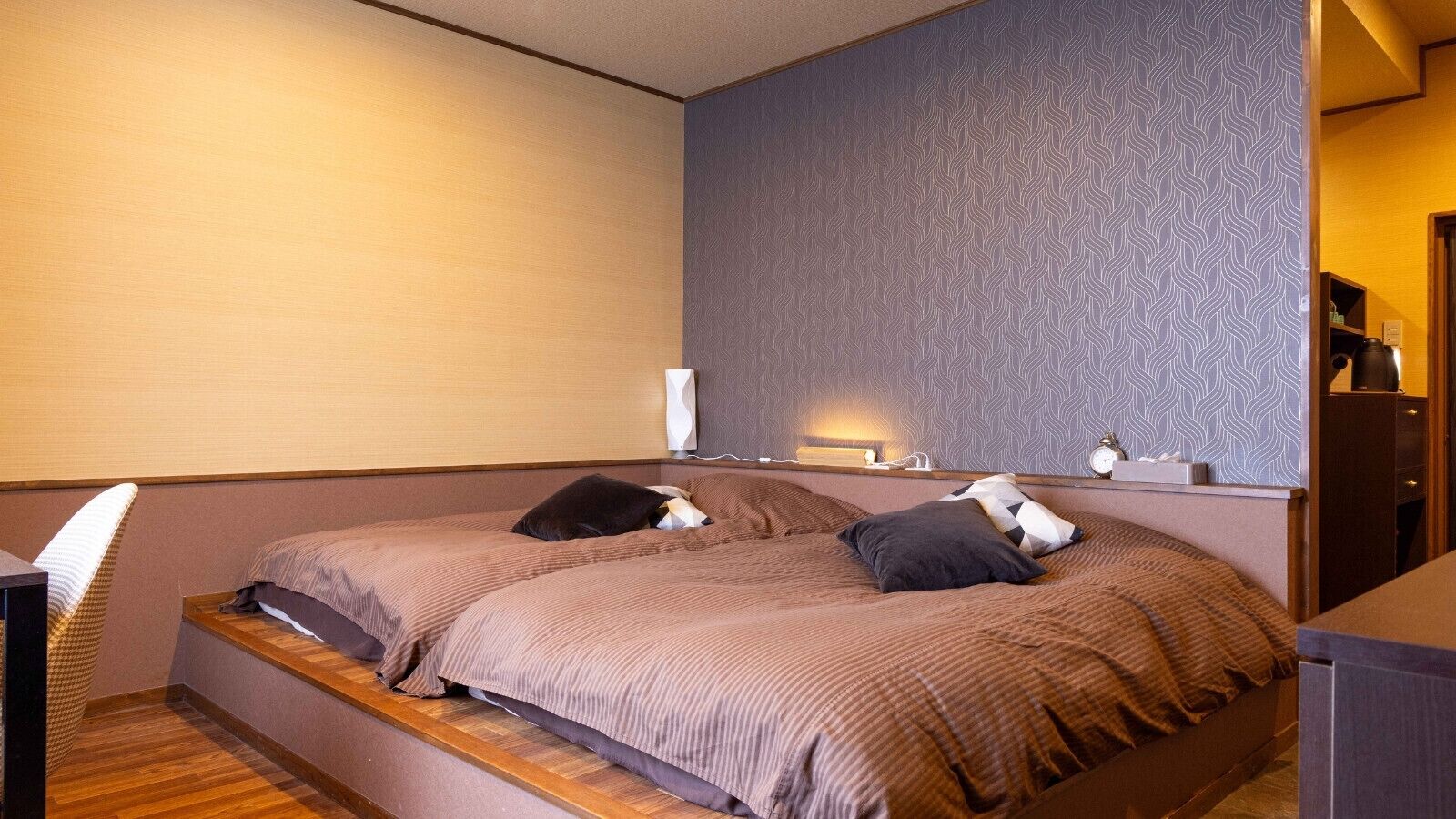 A bedroom with two beds, a wooden bench, a desk, and a wall-mounted shelf with a lamp.