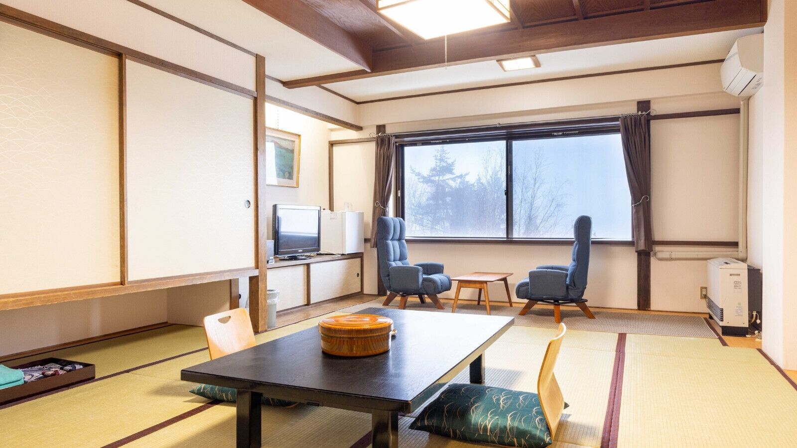 A room with tatami flooring, a low table with a wooden container, two blue chairs, a television, and a large window with curtains.