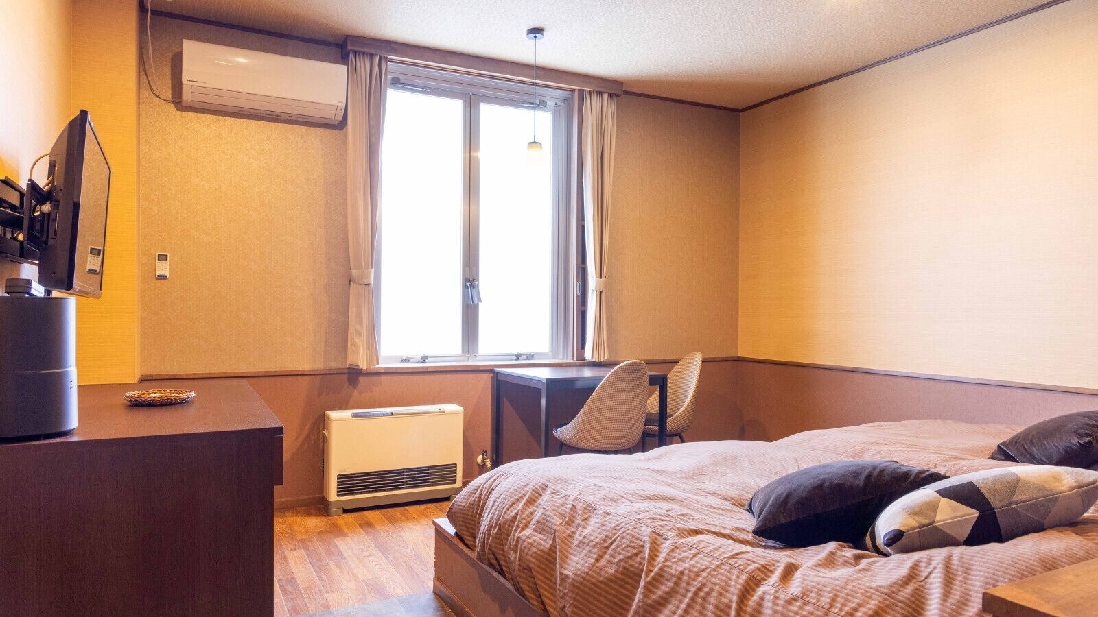 A hotel room with a bed, a desk, a chair, an air conditioning unit, and a window.