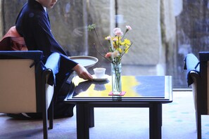 A person in traditional attire sitting at a table with a cup and saucer, a vase with flowers, and a window in the background. - Shisuitei (Numata)