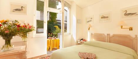 Un dormitorio con una cama grande, una mesita de noche con un florero y vistas al jardín a través de una puerta abierta.