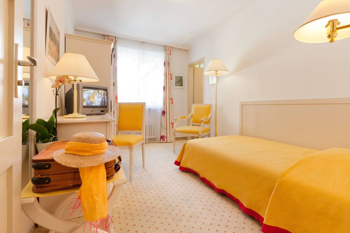 A hotel room with a bed, a desk with a television, two chairs, and a suitcase.