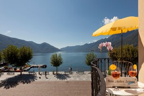 Una terraza junto al lago con un paraguas amarillo, una mesa con dos copas de vino y un periódico titulado "Meine Zeitung".
