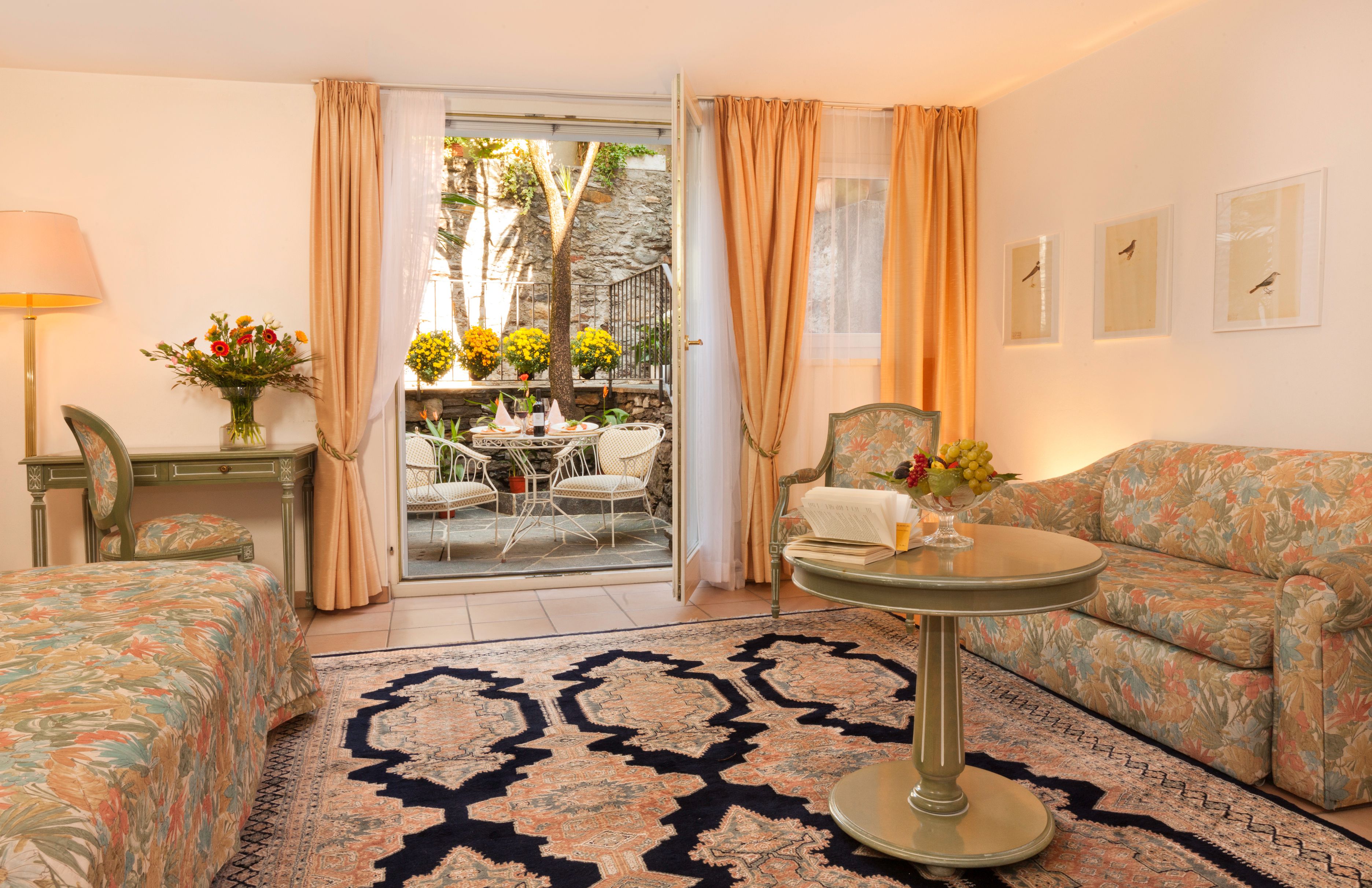 A hotel room with a bed, a sofa, a dining table with chairs, a small balcony with a table and chairs, and a view of a garden.