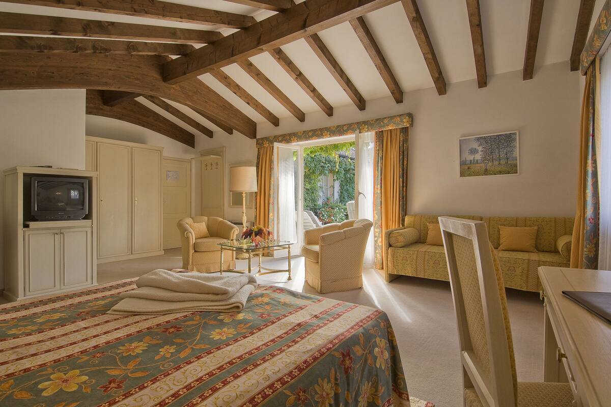 A bedroom with a large bed, a television, a sofa, and a chair. There is a window with curtains and a painting on the wall.