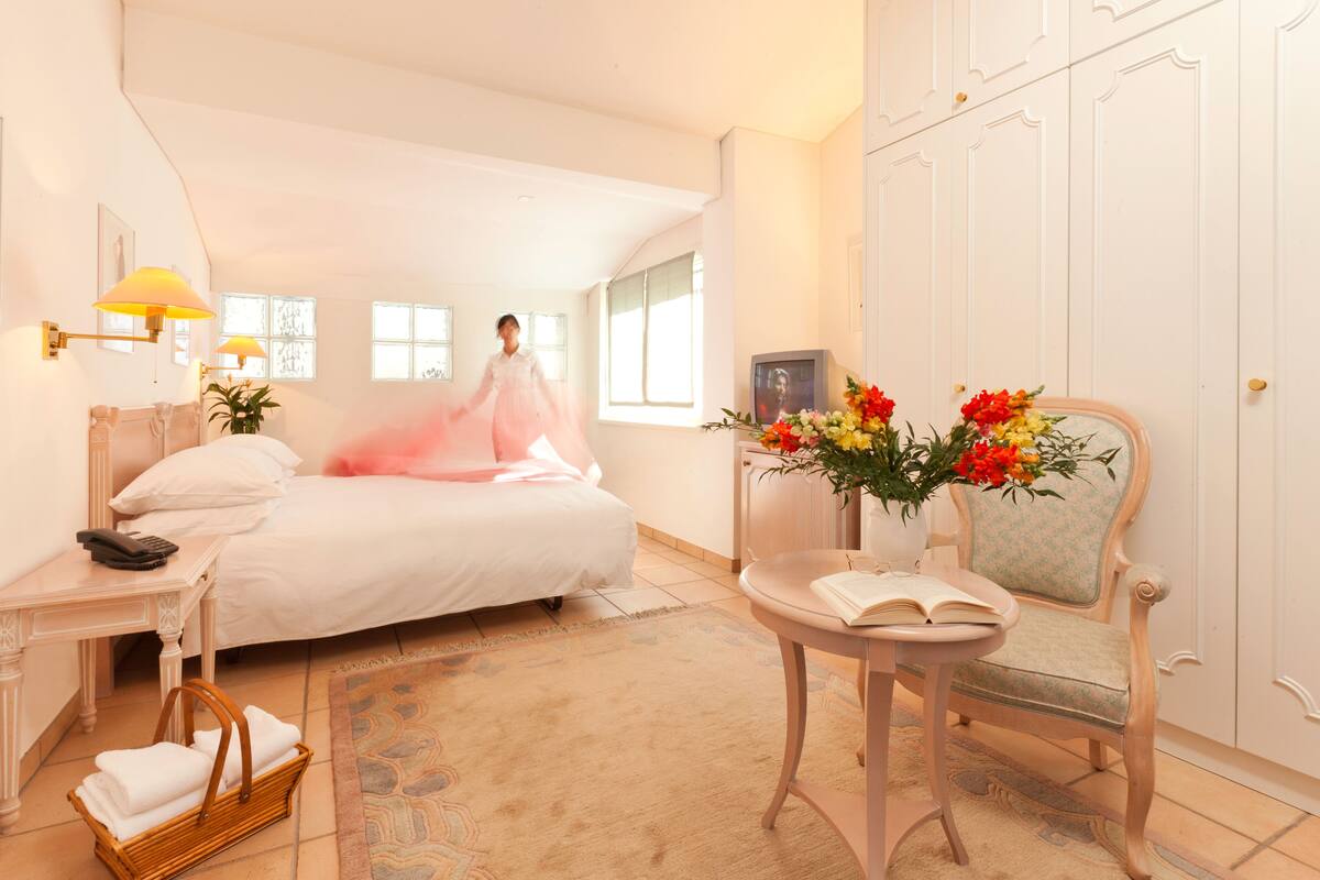A hotel room with a bed, a chair, a table with a book, a basket of towels, and a vase of flowers.