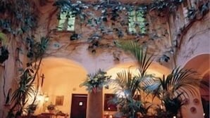 Ein Restaurant mit hohen Decken und einer Glasüberdachung, durch die natürliches Licht einflutet. Die Tische sind mit weißen Tischdecken gedeckt, und es befinden sich mehrere Blumentöpfe im Raum.
