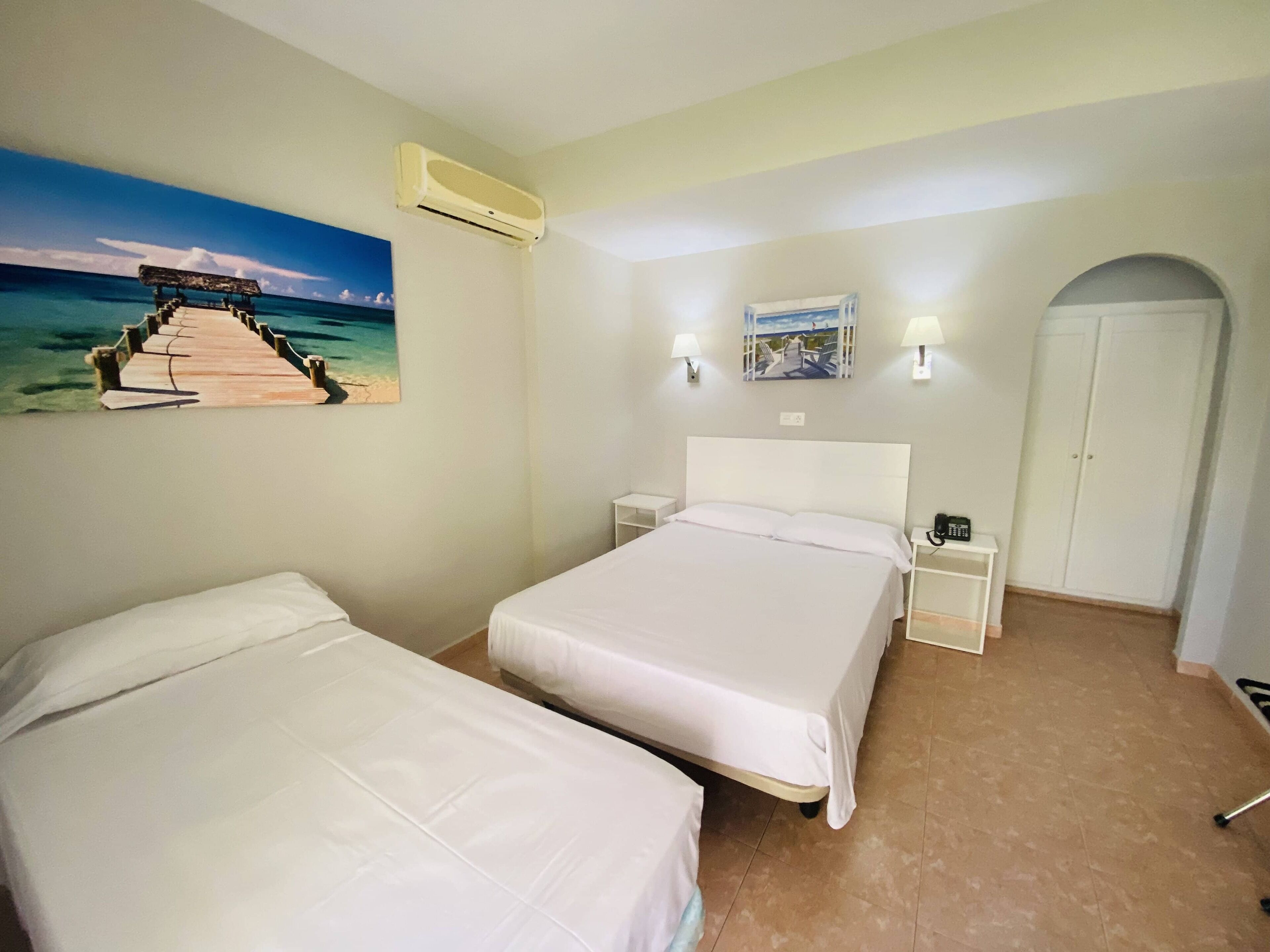 Habitación de hotel con dos camas, un cuadro de una escena de playa, aire acondicionado, una mesita de noche con un teléfono y una puerta.