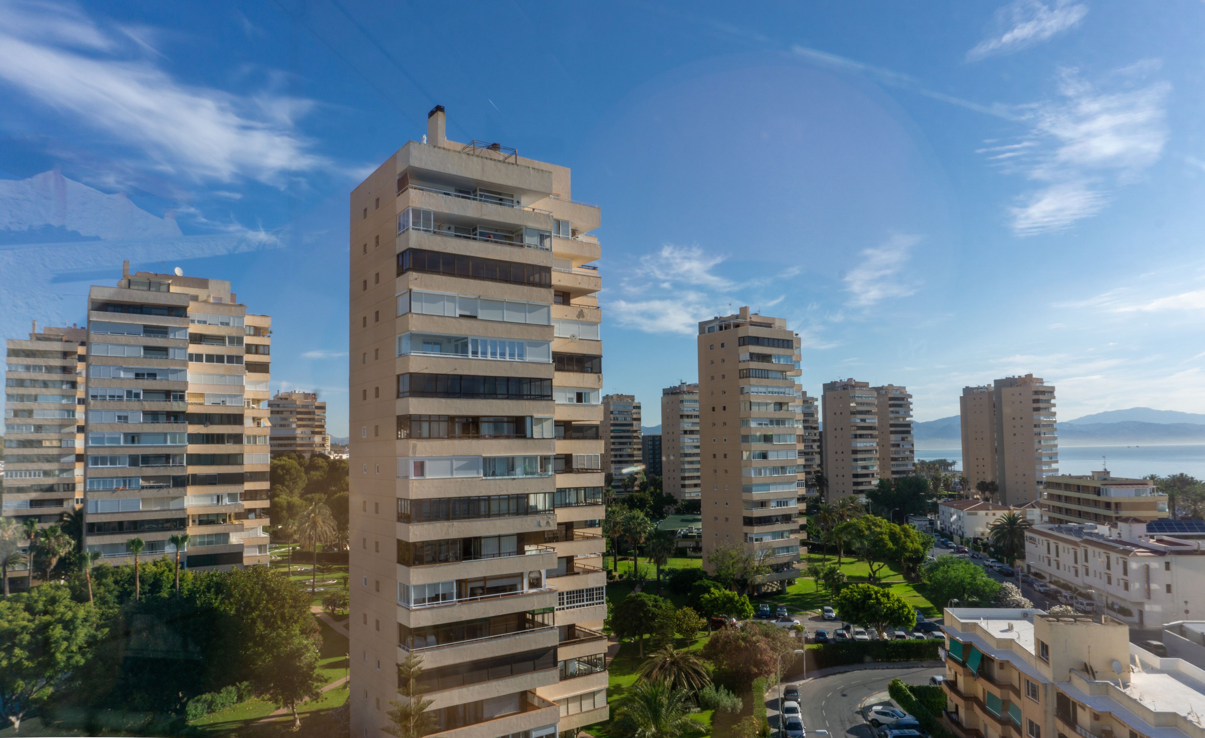 A high-rise residential building with multiple balconies, surrounded by greenery and other buildings.