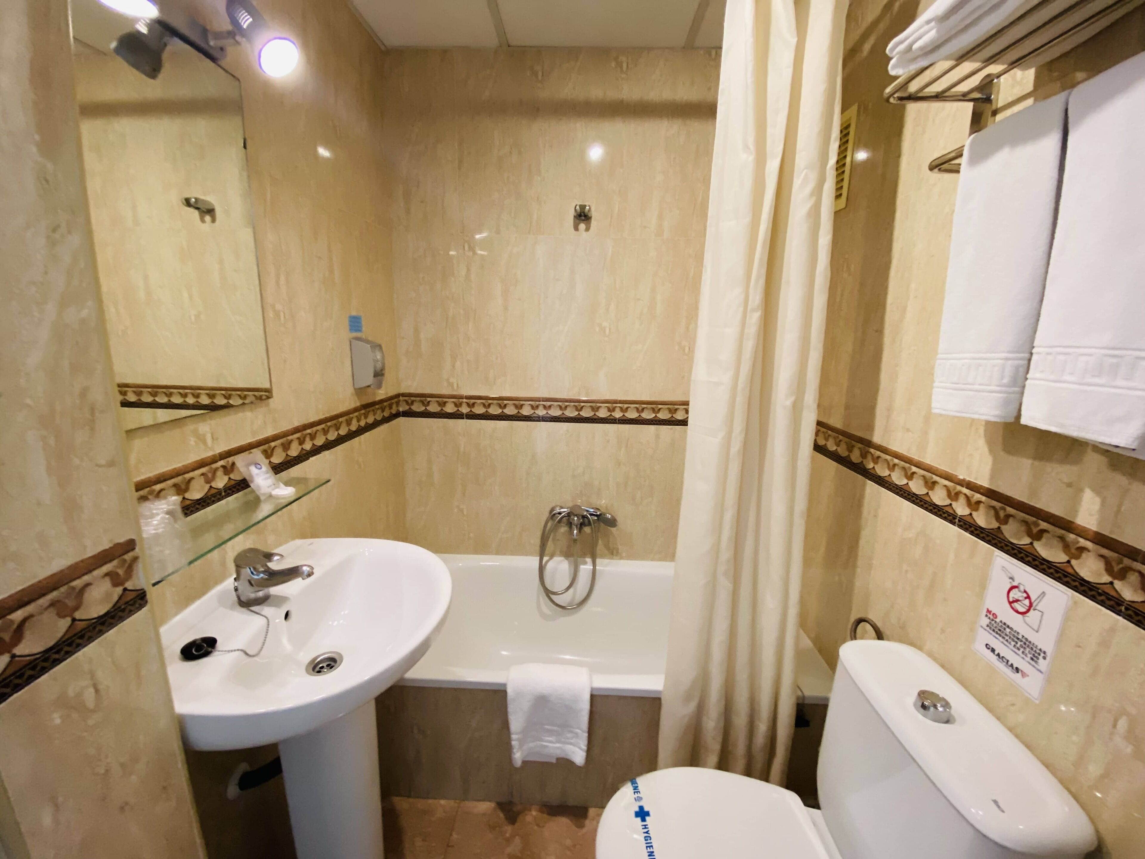 Un baño con lavamanos, bañera y retrete. Hay un perchero con toallas, un estante de vidrio con artículos de tocador y un letrero que indica que no se permite fumar.