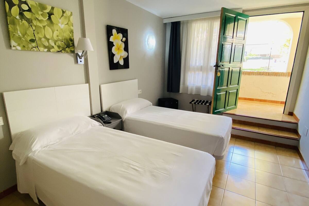 Een hotelkamer met twee bedden, een nachtkastje, een lamp, een schilderij en uitzicht op buiten.