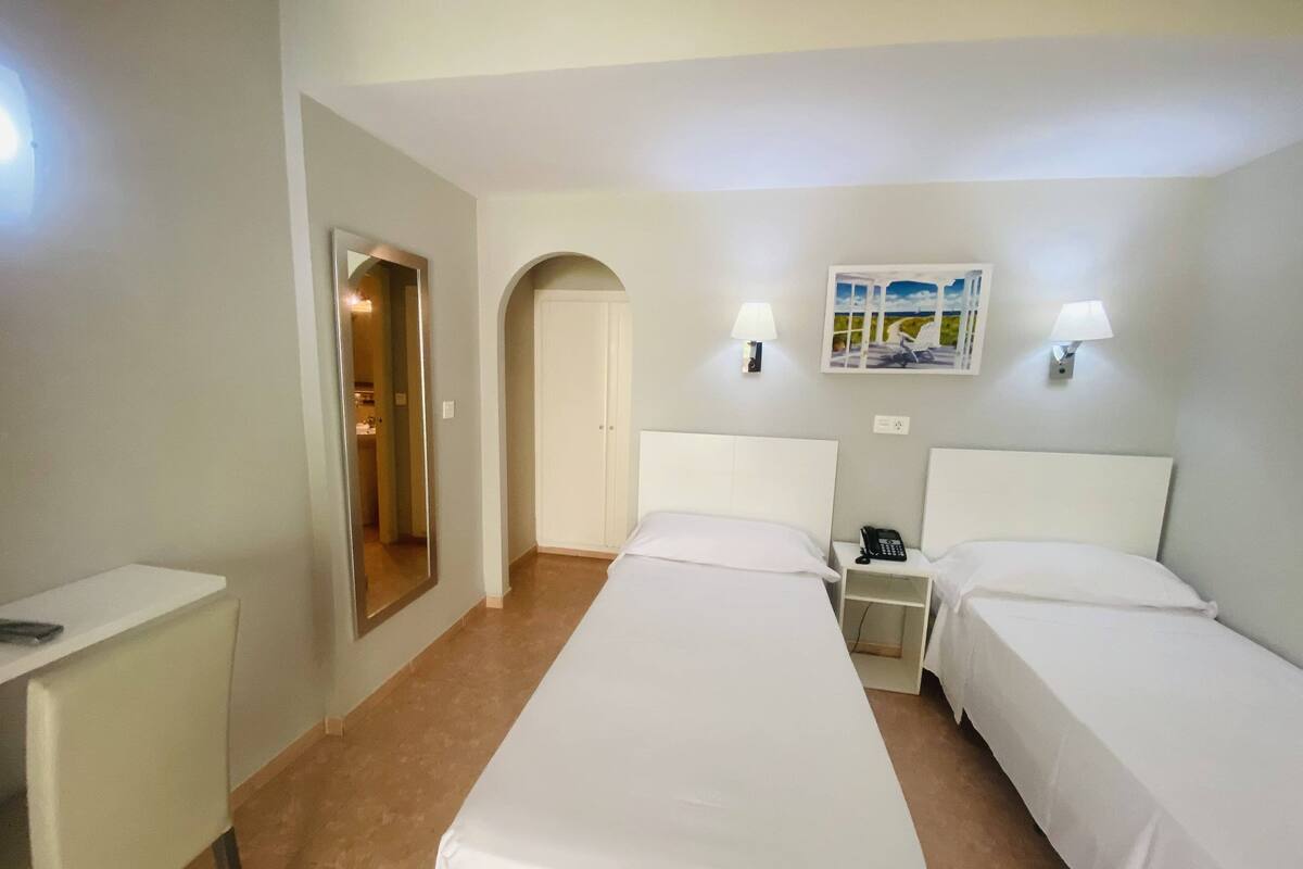 Een hotelkamer met twee eenpersoonsbedden, een bureau, een stoel, een nachtkastje met een telefoon en een deur die naar een andere kamer leidt.