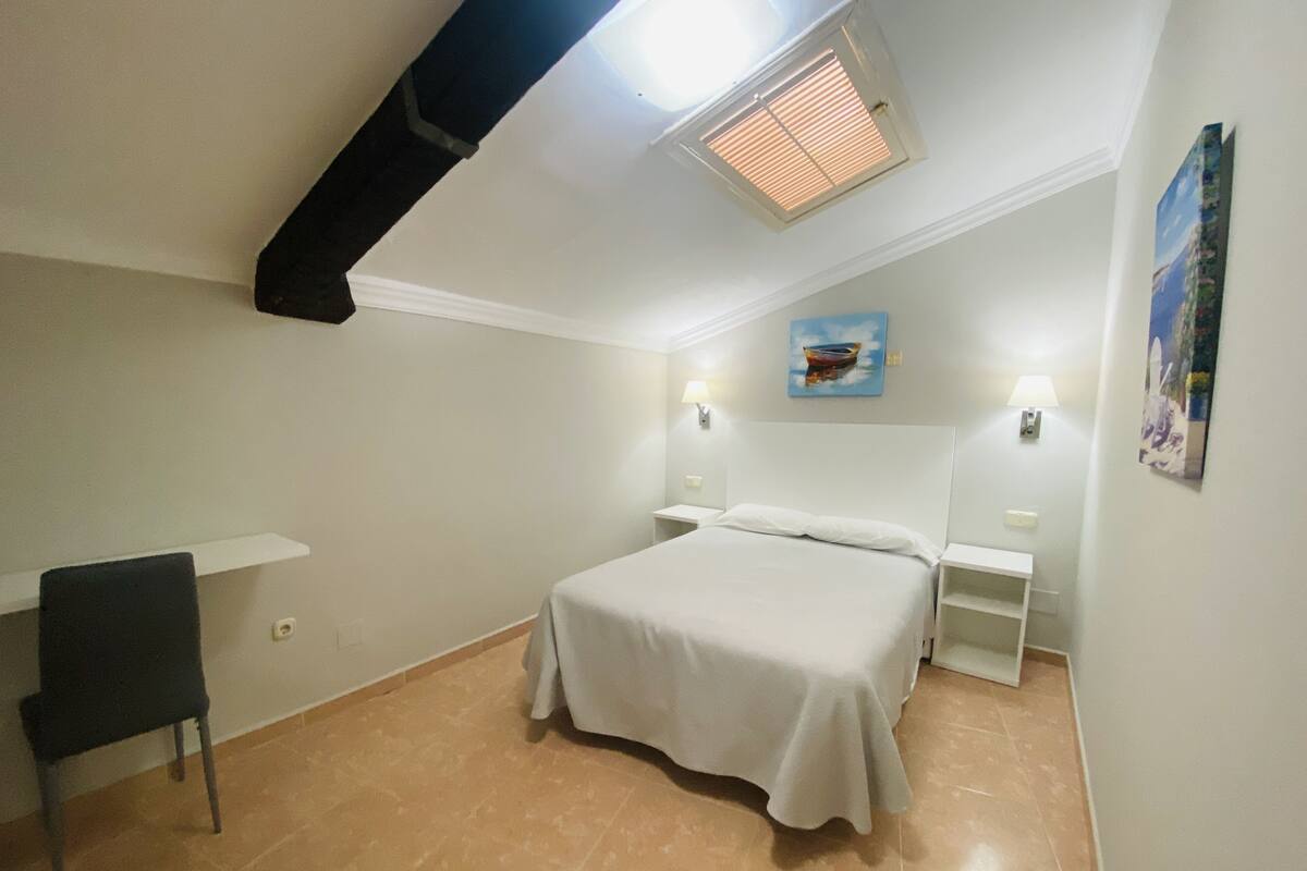 Een slaapkamer met een bed, een stoel, een nachtkastje, een wandlamp en een ventilatie rooster.