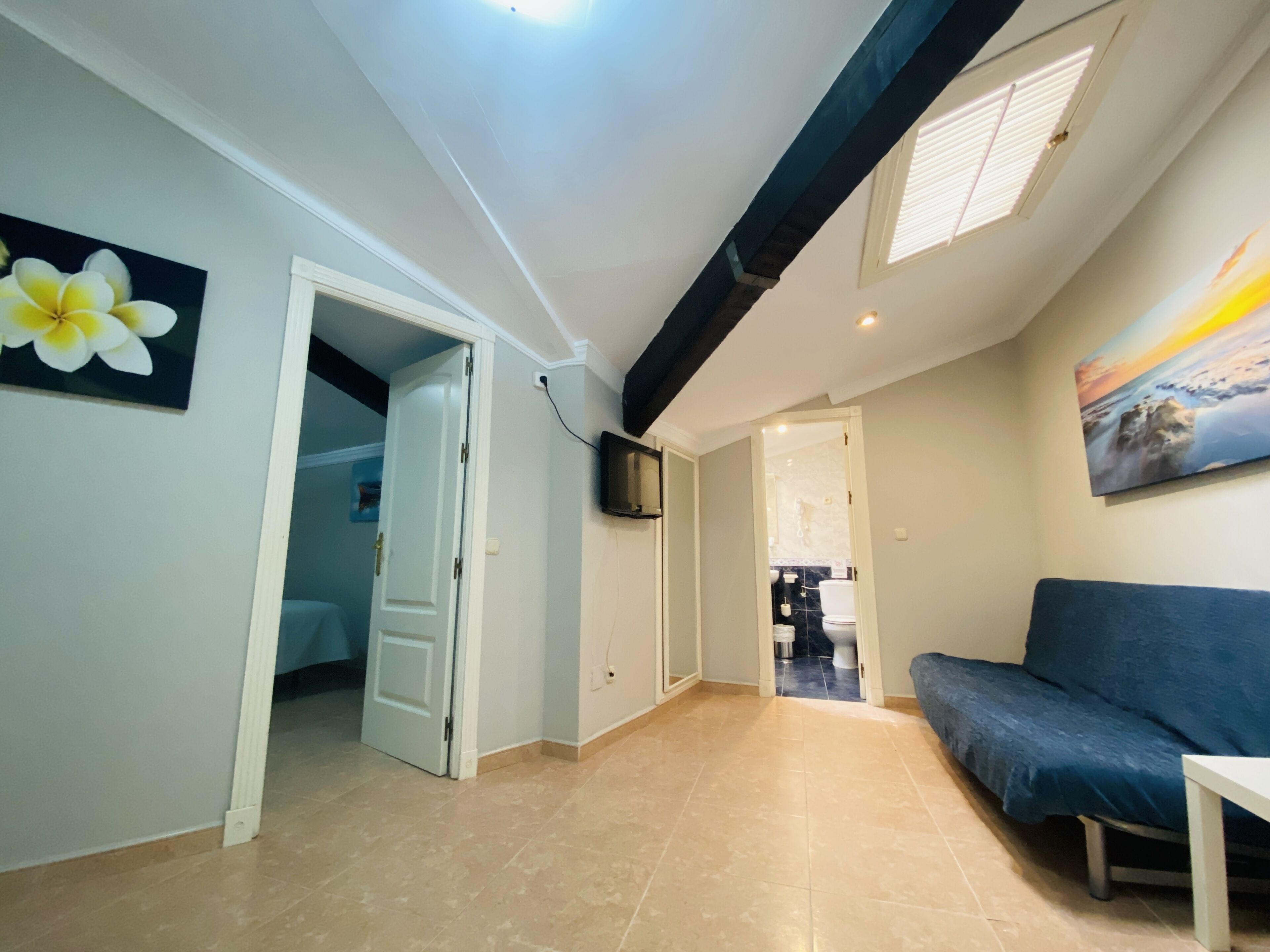 A spacious living area with a blue sofa, a door leading to a bathroom, and a TV mounted on the wall.