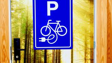A blue parking sign with a bicycle and plug symbol, set against a forest backdrop.